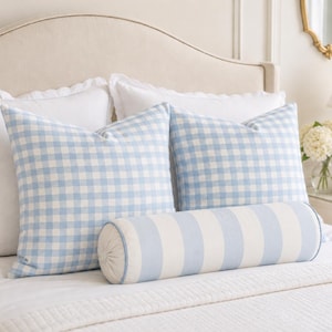 May include: Two square throw pillows featuring a light blue and white gingham check pattern, accompanied by a cylindrical bolster pillow with wide light blue and white vertical stripes and light blue piping along the circular end edges.
