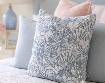 May include: A square throw pillow featuring a repeating blue and white seashell pattern on a white background. The pillow is positioned on a bed with other pillows and bedding. The text Dream Decor Ideas is visible in the bottom right corner.