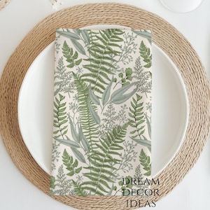 May include: A folded cloth napkin with a fern leaf pattern rests on a white plate, set on a woven placemat. The napkin features green and gray fern illustrations on a cream background. Silverware and a water glass are also visible.