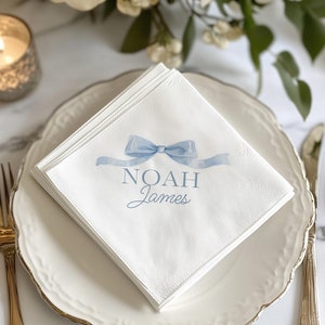 May include: White paper napkins with a blue ribbon bow and the text "NOAH James" printed on them. The napkins are folded and placed on a white plate with a gold rim.