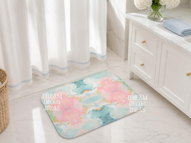 May include: A rectangular bath mat featuring a soft, watercolor-style abstract pattern in shades of pastel pink, teal, and white. The text DREAM DECOR IDEAS is printed in white on the left and right sides of the mat, which has rounded corners.