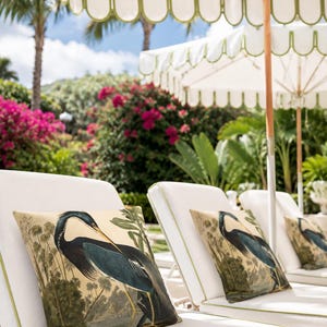 May include: White chaise lounges with green trim and decorative pillows featuring a blue heron illustration. The scene is set outdoors with white umbrellas, lush greenery, and pink flowers in the background.