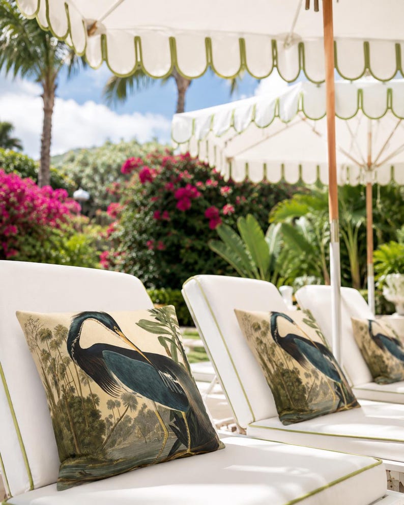 May include: White chaise lounges with green trim and decorative pillows featuring a blue heron illustration. The scene is set outdoors with white umbrellas, lush greenery, and pink flowers in the background.