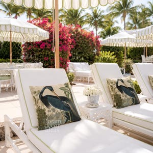 May include: White chaise lounges with green trim and matching pillows featuring a blue heron design. White umbrellas with green scalloped edges provide shade. The scene is set outdoors with palm trees and flowering bushes.