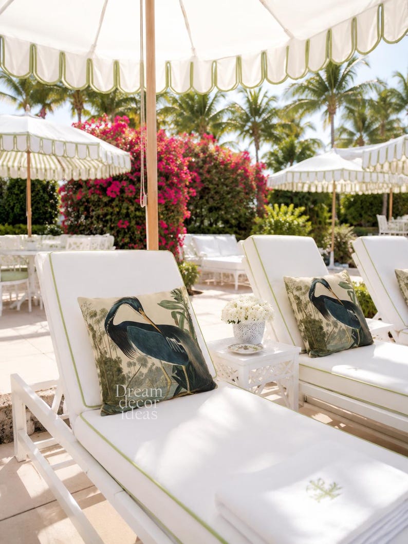 May include: White chaise lounges with green trim and matching pillows featuring a blue heron design. White umbrellas with green scalloped edges provide shade. The scene is set outdoors with palm trees and flowering bushes.