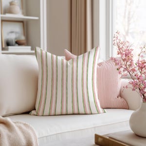 May include: A decorative pillow with a white background and vertical stripes in pink and green. The pillow is on a white sofa with other pillows and a blanket. A vase of pink flowers is in the background.
