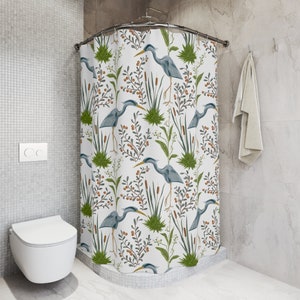 May include: A white shower curtain with a blue heron and green foliage pattern. The shower curtain is hung on a curved shower rod.