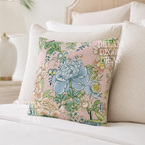 May include: A square throw pillow featuring a light pink background with a floral pattern of blue, yellow, and green flowers and leaves. The text DREAM DECOR IDEAS is printed in white on the front of the pillow. The pillow is placed on a bed.