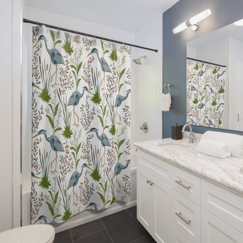 May include: A white shower curtain with a repeating pattern of blue herons, green reeds, and brown berries. The herons are standing in a marsh-like setting.