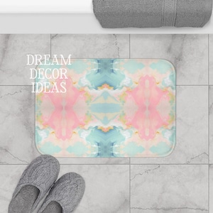 May include: A rectangular bath mat featuring a symmetrical, marbled pattern in soft shades of pastel pink, light blue, and cream. The text DREAM DECOR IDEAS is printed in white on the left side of the mat, which has rounded corners.