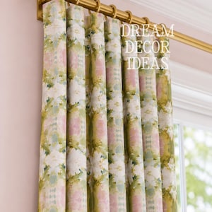 May include: A floor-length curtain featuring a watercolor-style floral pattern in shades of sage green, soft pink, and white. The fabric hangs from a metallic gold curtain rod with matching rings. The text DREAM DECOR IDEAS is visible.