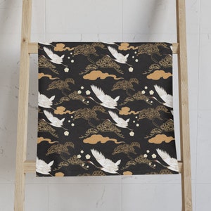 May include: A black towel with a repeating pattern of white cranes in flight, gold clouds, and stylized trees. The towel is hanging on a wooden ladder-style rack. The design has a Japanese aesthetic.