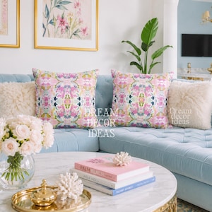 May include: Two square throw pillows featuring a symmetrical, abstract floral pattern in shades of pink, green, and white. The pillows are positioned on a light blue tufted sofa, flanked by two cream-colored, textured faux fur throw pillows.