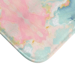May include: A rectangular bath mat featuring a soft, textured surface with an abstract marble pattern in shades of pastel pink, light blue, and white, accented with subtle swirls of yellow and gold throughout the marbled design.