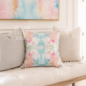 May include: A square throw pillow featuring a watercolor-style pattern in shades of pink, blue, and yellow, flanked by two solid beige textured pillows. The text DREAM DECOR IDEAS is overlaid on the image in white, semi-transparent lettering.