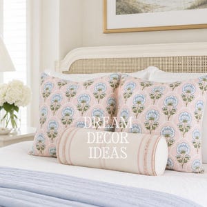 May include: Two square throw pillows featuring a light pink background with a repeating blue and green floral pattern, accompanied by a cylindrical bolster pillow in a solid cream color with subtle vertical stripes and the text DREAM DECOR IDEAS.