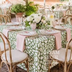 May include: A square table set for dining with a green and white lattice patterned tablecloth, light pink cloth napkins, white plates with floral accents, pink textured glassware, and a central floral arrangement of white hydrangeas.