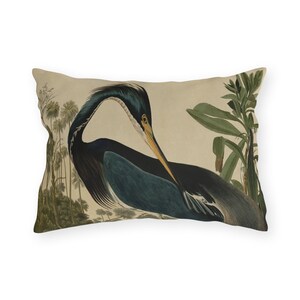 May include: Rectangular pillow featuring a vintage illustration of a heron. The bird has dark blue and black feathers, a yellow beak, and is set against a beige background with green foliage and trees. The pillow has a neutral color palette.