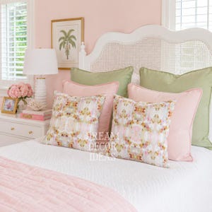 May include: A white cane headboard with a bed featuring a white quilted coverlet, two solid sage green square pillows, two solid light pink square pillows, and two square pillows with a multicolored abstract floral pattern in pink, green, and white.