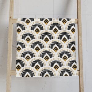 May include: A white towel with a repeating Art Deco fan pattern in black and gold. The towel is draped over a wooden ladder, showcasing the geometric design. The pattern features semi-circular shapes with a central arrow design.