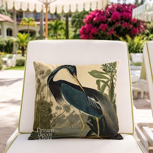 May include: A decorative pillow with a vintage illustration of a blue heron in a tropical setting. The pillow is on a white outdoor chair with green trim. The words "Dream decor ideas" are printed on the pillow.