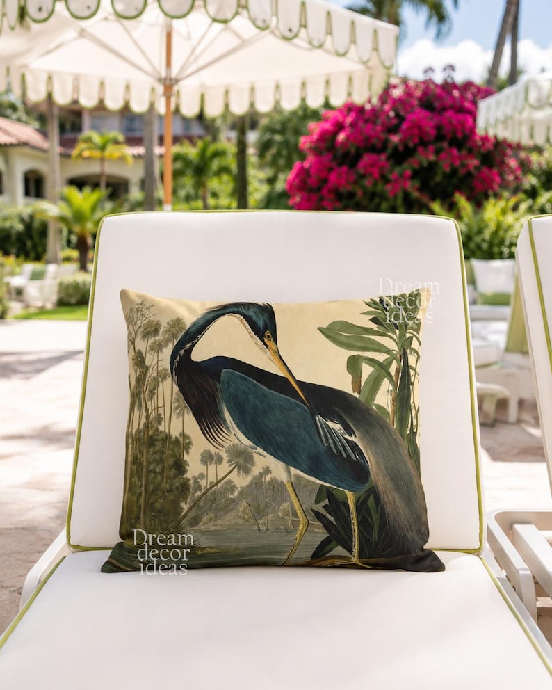 May include: A decorative pillow with a vintage illustration of a blue heron in a tropical setting. The pillow is on a white outdoor chair with green trim. The words "Dream decor ideas" are printed on the pillow.