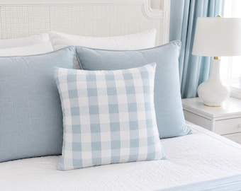 May include: A square throw pillow featuring a light blue and white gingham check pattern, positioned in front of two larger solid light blue pillows on a white bedspread. The pillow has a clean, simple design with a visible fabric texture throughout.