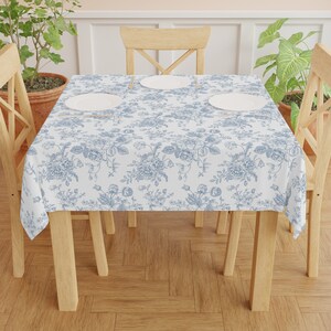 May include: A white tablecloth with a blue floral pattern, draped over a wooden table. The floral design features detailed illustrations of roses and other flowers. Three white plates are set on the table.