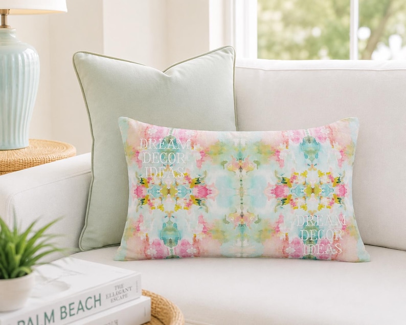 May include: A rectangular throw pillow featuring a vibrant, symmetrical watercolor pattern in shades of pink, yellow, and light blue. The text "DREAM DECOR IDEAS" is printed in white on the left and right sides of the pillow surface.