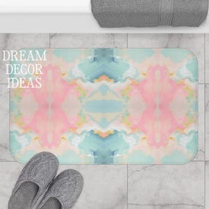 May include: A rectangular bath mat featuring a symmetrical, marbled pattern in soft shades of pink, light blue, and cream. The design displays fluid, cloud-like swirls across the surface. The text DREAM DECOR IDEAS is printed in white.