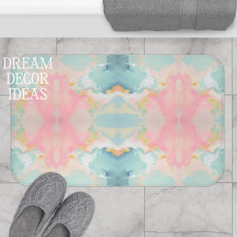May include: A rectangular bath mat featuring a symmetrical, marbled pattern in soft shades of pink, light blue, and cream. The design displays fluid, cloud-like swirls across the surface. The text DREAM DECOR IDEAS is printed in white.