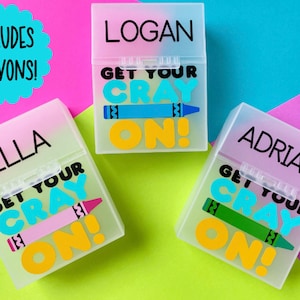 May include: Three personalized crayon boxes with the names "Logan", "Ella", and "Adrian" printed on them. Each box has the phrase "Get Your Cray On!" and a crayon graphic. The boxes are on a colorful background with the text "Includes Crayons!"