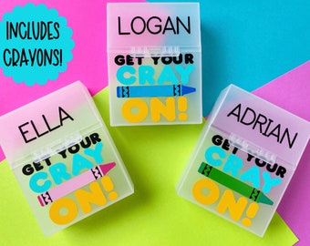 Op de afbeelding: Drie gepersonaliseerde kleurpotloodoosjes met de namen "Logan", "Ella" en "Adrian". Elke doos heeft de tekst "Get Your Cray On!" en een kleurpotloodafbeelding. De achtergrond is kleurrijk, met de tekst "Includes Crayons!"