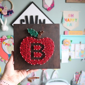 May include: A red apple with a green stem and leaf is made with string art on a dark brown wooden square. The letter "B" is woven into the apple.