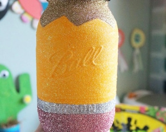 May include: A glass mason jar decorated to look like a pencil. The jar is covered in yellow glitter with a silver glitter band and a pink glitter eraser. The top of the jar is black glitter.