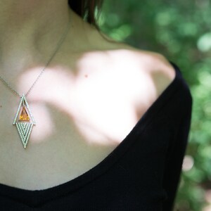 May include: A silver necklace with a geometric pendant featuring a central orange gemstone and surrounding white gemstones. The necklace is worn against a black top, with a blurred green background.