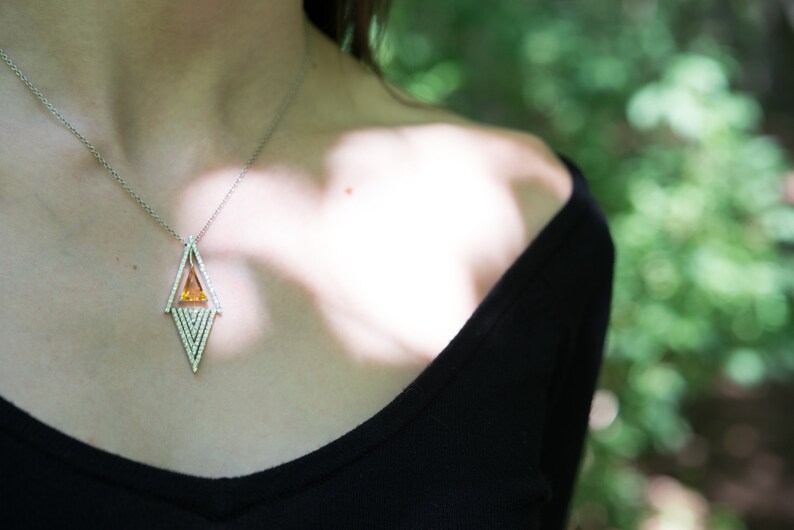 May include: A silver necklace with a geometric pendant featuring a central orange gemstone and surrounding white gemstones. The necklace is worn against a black top, with a blurred green background.