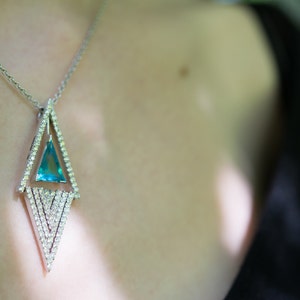 May include: Silver necklace with a geometric pendant. The pendant features a triangular, light blue gemstone set within a diamond-studded frame. The necklace is worn against a dark background, highlighting the jewelry's sparkle and design.