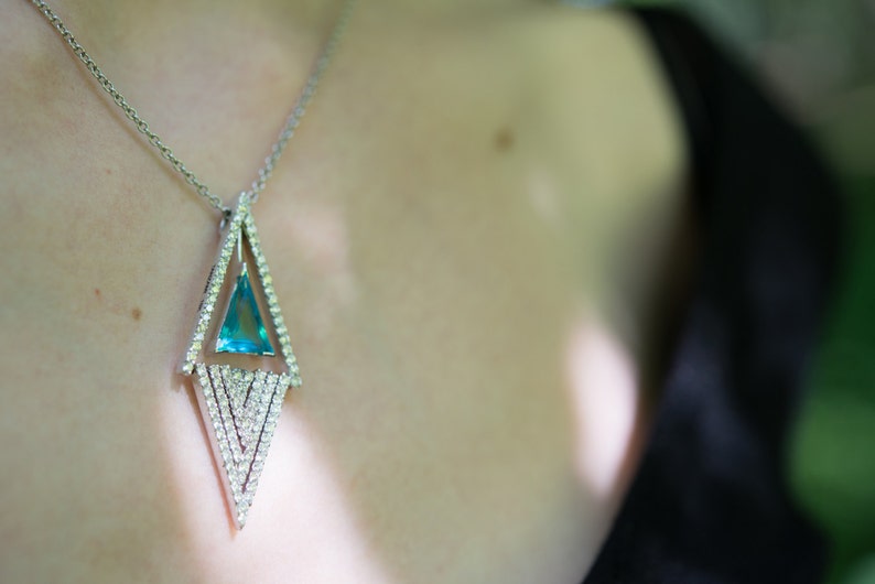 May include: Silver necklace with a geometric pendant. The pendant features a triangular, light blue gemstone set within a diamond-studded frame. The necklace is worn against a dark background, highlighting the jewelry's sparkle and design.