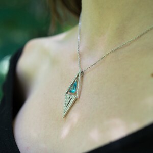 May include: A silver necklace with a geometric pendant. The pendant features a light blue gemstone set within a diamond-shaped frame, accented with small, sparkling stones. The necklace is worn against a dark background.