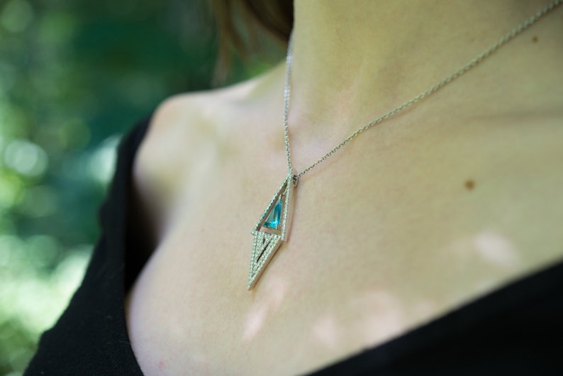 May include: A silver necklace with a geometric pendant. The pendant features a light blue gemstone set within a diamond-shaped frame, accented with small, sparkling stones. The necklace is worn against a dark background.