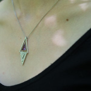 May include: A silver necklace with a geometric pendant. The pendant features a purple gemstone set within a diamond-shaped frame, accented with small, clear stones. The necklace is worn against a black top.