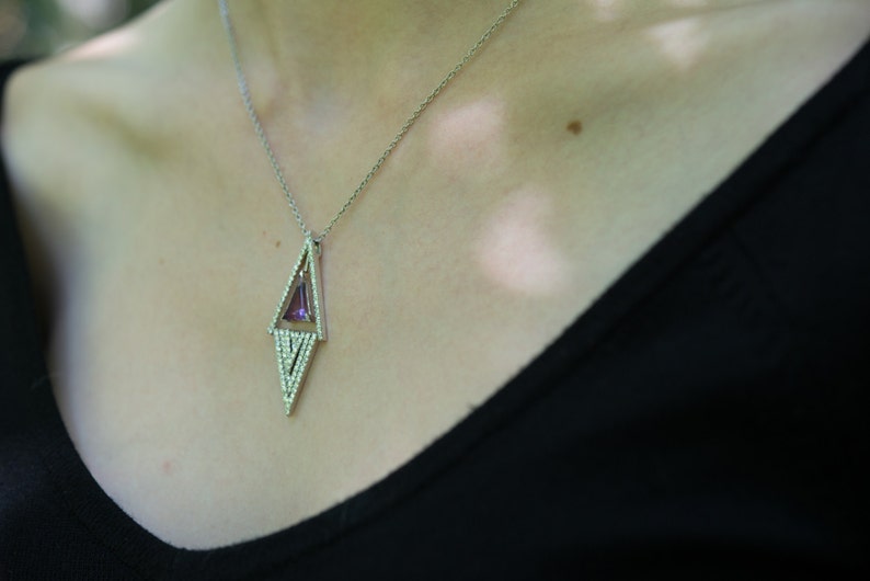 May include: A silver necklace with a geometric pendant. The pendant features a purple gemstone set within a diamond-shaped frame, accented with small, clear stones. The necklace is worn against a black top.