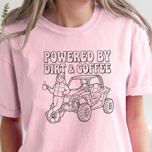 May include: Light pink t-shirt with the text "POWERED BY DIRT & COFFEE" above a black and white graphic of a person holding a coffee mug next to an off-road vehicle. The shirt has rolled-up sleeves.