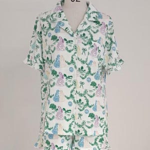 May include: White pajama set with a whimsical print featuring figures in gowns, trees, and bows. The short-sleeved top and shorts have a ruffled hem. The print includes green, blue, purple, and yellow accents.