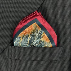 May include: A burgundy pocket square with a dark green and orange floral design, folded and placed in a dark brown suit pocket. The pocket square has a dark green border and a burgundy edge.