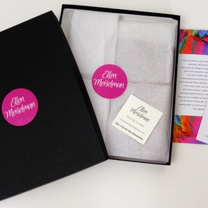 May include: Black gift box with a pink circular sticker that reads "Ellen Meiselman". Inside the box is white tissue paper and a colorful scarf. A small card and a thank you note are also visible.