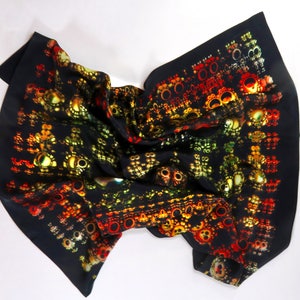 Op de afbeelding: Een zwarte zijden sjaal met een herhalend patroon van kleurrijke, abstracte vormen in rood, geel en groen. De sjaal is gevouwen en gedrapeerd, wat het ingewikkelde ontwerp benadrukt. Modeaccessoire.