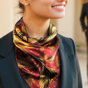 May include: A square scarf featuring a deep burgundy background with a prominent gold and black leaf pattern and small white square accents. The scarf is tied around the neck and worn over a dark blazer with a single visible button at the bottom.