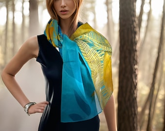 May include: A woman wearing a dark blue sleeveless dress and a colorful scarf in a forest setting. The scarf features a blue and yellow design with leaf patterns. A silver watch is on the woman's wrist.